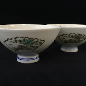 May include: Two white ceramic bowls with a flared rim and a pedestal base. Each bowl features a hand-painted fan-shaped graphic on the exterior, depicting a landscape scene with green trees and brown structures, accented by blue rings on the base.