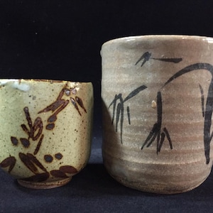 May include: Two handmade ceramic teacups. The smaller cup is light green with brown floral designs. The larger cup is tan with black bamboo-style brushstrokes. Both cups have a rustic, textured appearance.