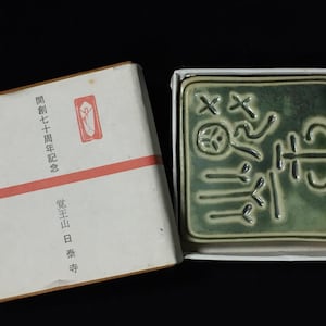 以下が含まれることがあります： 白い箱に入った、浮き彫りの日本語の文字が入った緑色のセラミックスタンプ。箱は開いており、スタンプが見えています。隣には日本語のテキストが入った白と赤の箱があります。