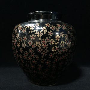 Puede incluir: Un jarrón de cerámica negra con cuerpo redondeado y cuello corto, que presenta un patrón repetitivo de pequeñas flores de color dorado con diminutos centros rojos distribuidos por toda la superficie exterior.