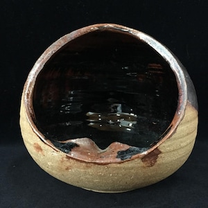 May include: A spherical ceramic bowl with a wide, angled opening. The exterior features a textured, light tan finish with horizontal ridges, while the interior is coated in a smooth, glossy black glaze that spills over the rim in dark brown.