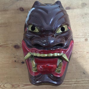 May include: A glossy brown ceramic mask featuring a stylized face with gold-colored eyes and sharp fangs. The mouth area is painted in a vibrant red, highlighting the gold teeth and tusks. The mask has a smooth, reflective finish throughout.