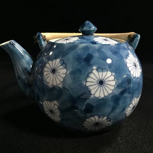 May include: Blue and white ceramic teapot with a floral pattern. The teapot has a rounded body, a curved spout, and a lid with a small knob. The handle is made of a light-colored material. The floral design features white flowers with blue centers.