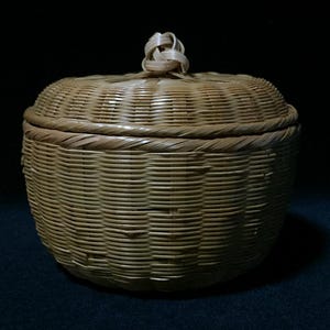 May include: A round, light brown woven basket with a matching lid. The basket features a tight, horizontal weave pattern and a decorative, knotted handle made of thin, flexible strips at the very center of the lid.