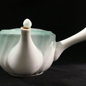 May include: A white and seafoam green ceramic teapot with a curved handle and a spout. The teapot has a lid with a small white knob. The body of the teapot has a textured, swirling design.