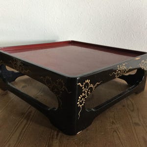 Puede incluir: Una bandeja de servicio cuadrada de perfil bajo con un exterior negro brillante y una superficie interior de color rojo intenso. Los lados negros presentan delicados patrones de volutas florales doradas pintadas a mano. La bandeja se apoya sobre cuatro patas arqueadas integradas.