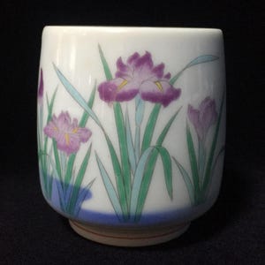 Może przedstawiać: Biały ceramiczny kubek do herbaty z ręcznie malowanym wzorem fioletowych kwiatów irysa z zielonymi liśćmi i łodygami. Podstawa kubka jest zaakcentowana poziomym niebieskim paskiem, a całość ma cylindryczny kształt z gładką, zaokrągloną krawędzią.