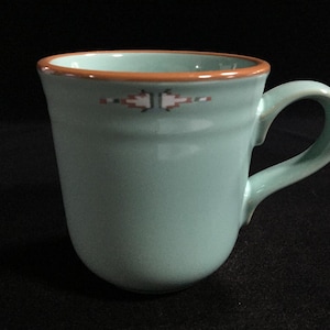 May include: A light mint green ceramic mug with a smooth, glossy finish and a curved handle. The rim is accented with a thin, contrasting terracotta-colored band. A small, geometric pattern in white, black, and terracotta sits below the rim.