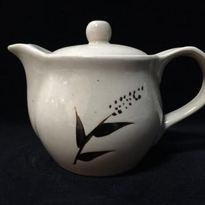 May include: Cream-coloured ceramic teapot with a lid and handle. The teapot has a black floral design on the side. The spout is on the left, and the handle is on the right. The lid has a small knob on top.