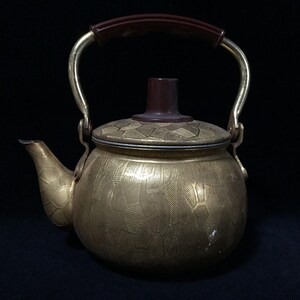 May include: A gold-toned metal tea kettle featuring a textured, geometric pattern across its surface. The kettle includes a curved spout, a dark brown knob on the lid, and a matching dark brown handle attached to a brass-colored metal frame.