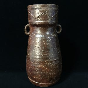 Può includere: Un vaso in ceramica marrone con una consistenza maculata e due piccole maniglie laterali. La sezione superiore presenta un collo largo con un motivo a tratteggio incrociato inciso attorno al bordo. Il corpo ha una forma affusolata con finitura screziata.