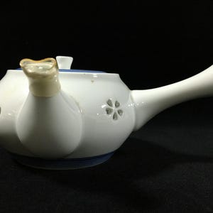 May include: White ceramic teapot with a blue rim and lid. The teapot features a long spout and a handle with a gold accent. A floral design is visible on the side of the teapot. The teapot is designed for brewing tea.