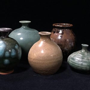 Peut inclure: Un ensemble de cinq vases en céramique de formes et tailles variées. Le groupe comprend un vase vert foncé avec des taches bleues, un vase vert clair lisse, un vase rond beige, un vase vernissé marron foncé et un petit vase vert côtelé plat.