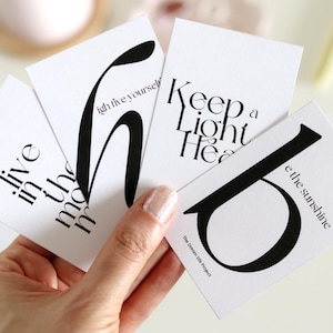May include: Four white cards with black text and graphic designs. The cards feature the phrases "live in the moment", "high five yourself", "keep a light heart", and "be the sunshine".