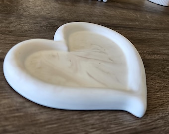 May include: A white heart-shaped trinket dish with a marbled interior. The dish is made of a solid material and sits on a wooden surface. The heart-shaped design is a decorative element.