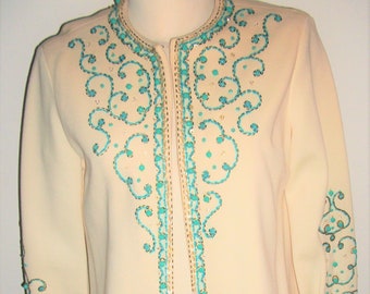 Puede incluir: Una chaqueta de color crema con cierre de cremallera y una intrincada pedrería y bordado de color turquesa y dorado. La pedrería y el bordado forman un patrón de espirales y bucles alrededor del cuello y por la parte delantera de la chaqueta.