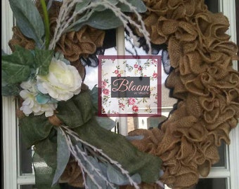 May include: A brown burlap wreath with a white and green floral arrangement and a green bow. The wreath has a black mesh backing and a sign that says "Bloom by Shayla".