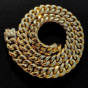 May include: A thick, polished gold-tone cuban link chain necklace arranged in a circular coil. The heavy metal links feature a high-shine reflective surface and a secure box clasp mechanism with a side safety latch, creating a bold, uniform appearance.