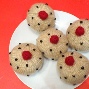 May include: Five knitted brown cupcakes with black dots and red centres on a white plate.