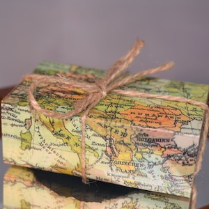 May include: A small, rectangular box wrapped in paper printed with a vintage world map design. The box is tied with twine, creating a rustic and travel-themed aesthetic. The map features shades of green, yellow, and brown.