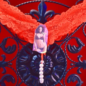 May include: Red lace lingerie with a tag featuring a woman wearing a black and white lingerie set. The tag is attached to a string of white pearls.