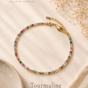 Puede incluir: Una pulsera delicada con pequeñas cuentas de turmalina facetadas y multicolores intercaladas con diminutas esferas de metal dorado. La pieza incluye un cierre de langosta dorado y una cadena extensora ajustable. Texto: ANNA LENA ARTISAN JEWELRY y Tourmaline.