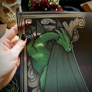 Puede incluir: Un drag&oacute;n verde con alas asoma desde detr&aacute;s de un marco decorativo. El drag&oacute;n est&aacute; dibujado en un estilo vintage con detalles intrincados.