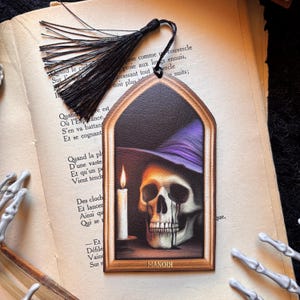 Peut inclure: Marque-page gothique avec un crâne portant un chapeau de sorcière violet, une bougie allumée et le mot "MANOIR". Le marque-page a une gland noir et est posé sur un livre ouvert avec du texte français.