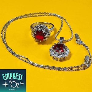 May include: A silver-toned jewellery set consisting of a ring and a pendant necklace. Both pieces feature a large, round, deep red centre stone surrounded by a halo of small, clear, white stones. The necklace has a thin, beaded metal chain.