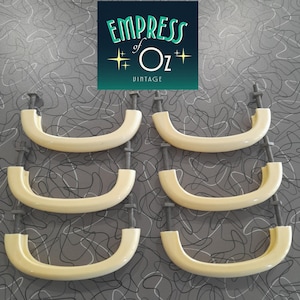 May include: Six off-white, semi-circular cabinet pulls arranged in two vertical columns. Each handle features a smooth, curved design with threaded metal bolts extending from both ends, including attached nuts for secure installation on furniture.