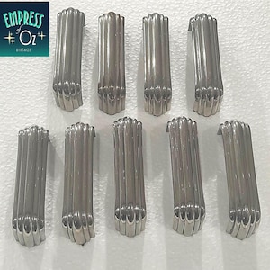May include: A set of nine silver-toned metal drawer pulls arranged in two rows. Each pull features a vertical, fluted, or ribbed design with a curved, arching profile. The metal has a smooth, reflective finish, and the handles are uniform in shape.
