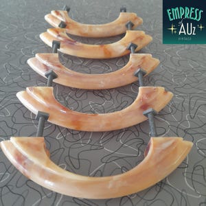May include: Five semi-circular drawer pulls made of a marbled, translucent peach and cream-coloured material. Each handle features two dark metal threaded bolts extending from the back for mounting, arranged in a vertical row on a grey surface.