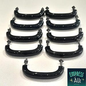 May include: A collection of nine black, curved drawer pulls arranged in rows. Each handle features a smooth, glossy finish and is attached to two metal mounting bolts with square-shaped hardware at the ends for secure installation on furniture.