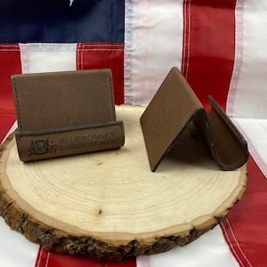 May include: Two brown leather phone stands with a black leather trim. The stand on the left has the text "Bluebonnet Laser Works" engraved on the front.