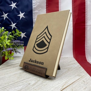 May include: A tan leather journal with a black Sergeant's rank insignia and the name "Jackson" printed on the front. The journal is standing upright on a wooden surface with a brown leather bookmark that reads "Bluebonnet Laser Works".
