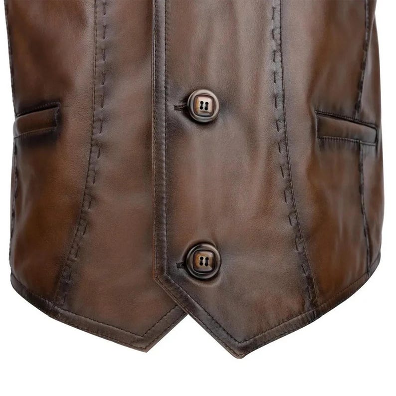 May include: A brown leather vest featuring a smooth finish, two dark brown buttons, and decorative whipstitch detailing along the seams. The garment includes two side pockets and a pointed hemline, showcasing a structured and tailored design.