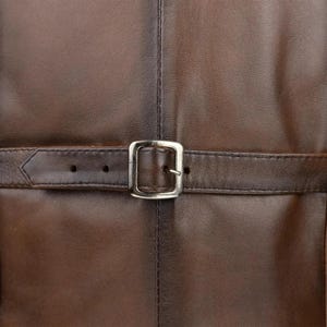 May include: A dark brown leather surface featuring a horizontal strap with a silver-toned metal buckle. The leather has a smooth texture with visible stitching running vertically down the center and along the edges of the strap.