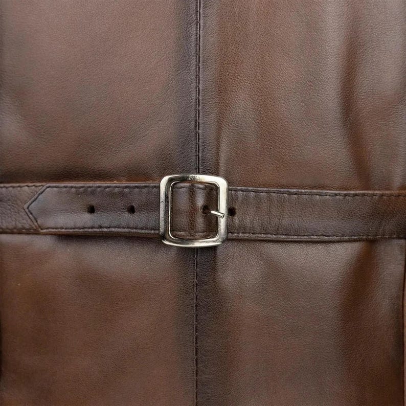 May include: A dark brown leather surface featuring a horizontal strap with a silver-toned metal buckle. The leather has a smooth texture with visible stitching running vertically down the center and along the edges of the strap.