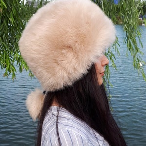 Peut inclure: Un chapeau en fausse fourrure beige de forme cylindrique avec une texture &eacute;paisse et pelucheuse. Le chapeau pr&eacute;sente un petit pompon en fausse fourrure beige assorti, attach&eacute; &agrave; l'arri&egrave;re par un fin cordon noir, offrant un aspect doux et arrondi.