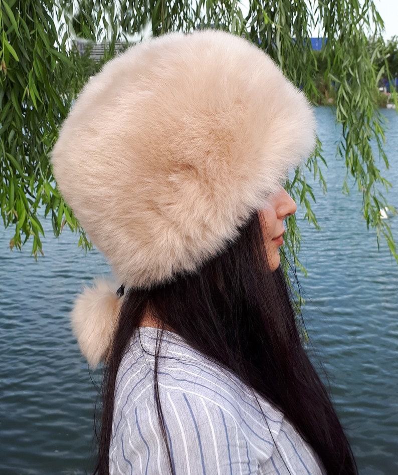 Peut inclure: Un chapeau en fausse fourrure beige de forme cylindrique avec une texture &eacute;paisse et pelucheuse. Le chapeau pr&eacute;sente un petit pompon en fausse fourrure beige assorti, attach&eacute; &agrave; l'arri&egrave;re par un fin cordon noir, offrant un aspect doux et arrondi.