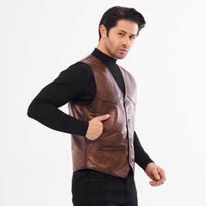 May include: A brown leather vest featuring a V-neckline, a front button closure with four dark buttons, and two side pockets. The vest has a smooth finish with visible stitching along the seams and a pointed hemline at the front waist area.