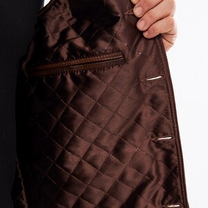 May include: A dark brown jacket interior featuring a quilted satin lining with a diamond pattern. The garment includes a horizontal welt pocket on the left side and a vertical row of buttonholes along the front edge, showcasing the construction.