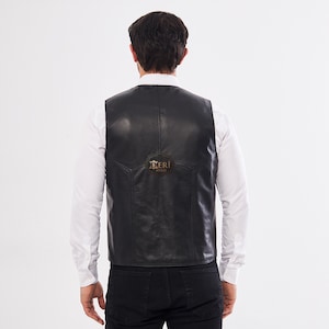 May include: A black leather vest worn over a white long-sleeved button-down shirt and black pants. The back of the vest features a smooth finish with visible seam detailing and a small, dark, circular logo patch with the text DERI SITESI.