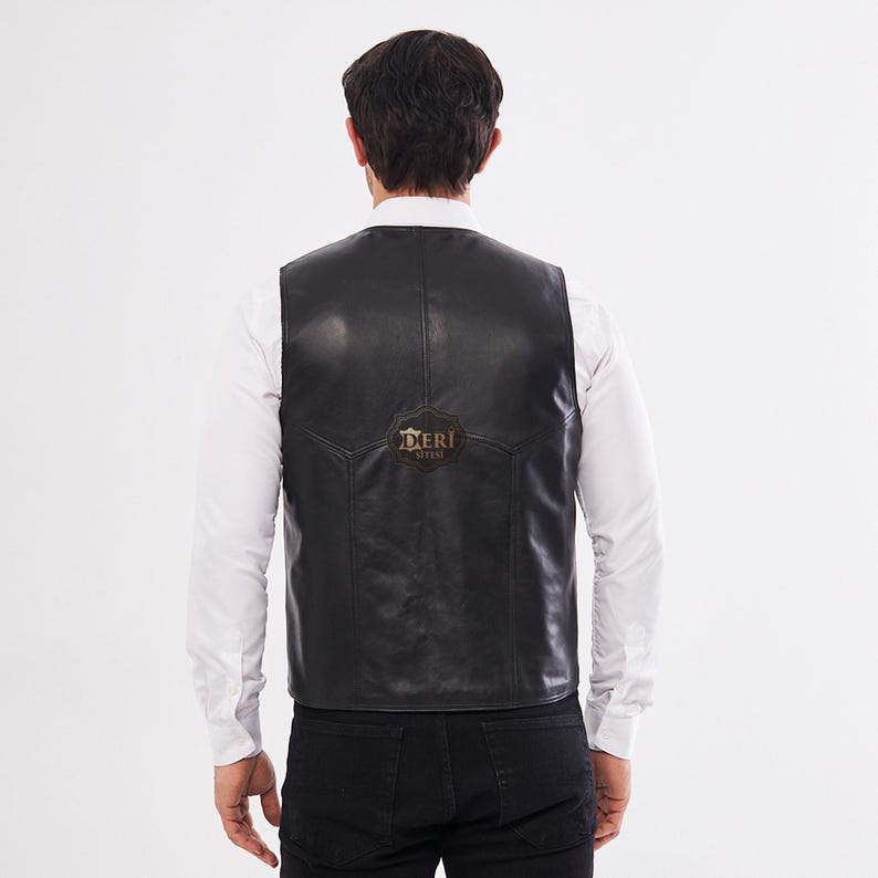 May include: A black leather vest worn over a white long-sleeved button-down shirt and black pants. The back of the vest features a smooth finish with visible seam detailing and a small, dark, circular logo patch with the text DERI SITESI.