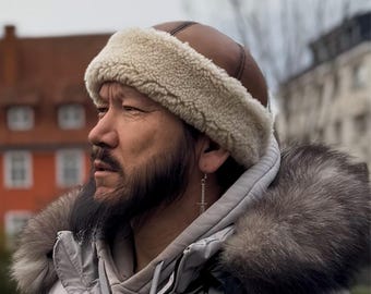 May include: A brown leather hat with a thick cream-colored shearling trim, worn with a light gray hooded jacket featuring a dark gray faux fur hood lining and a silver sword-shaped earring dangling from the earlobe.
