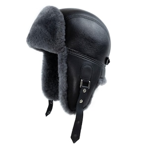 May include: A black leather trapper hat featuring a thick, dark gray shearling lining. The hat includes ear flaps that can be secured with black leather straps, silver-toned metal buckles, and snap buttons, providing full coverage for the head and ears.