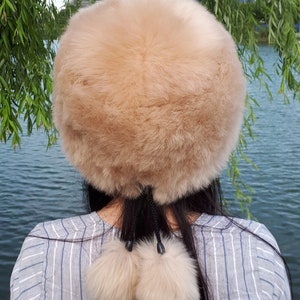 Peut inclure: Un bonnet rond en fausse fourrure beige, dot&eacute; d'une texture &eacute;paisse et douce. Le bonnet pr&eacute;sente deux pompons en fausse fourrure beige assortis, suspendus &agrave; de fines cordes noires &agrave; l'arri&egrave;re, formant un d&eacute;tail d&eacute;coratif sur le haut du dos.