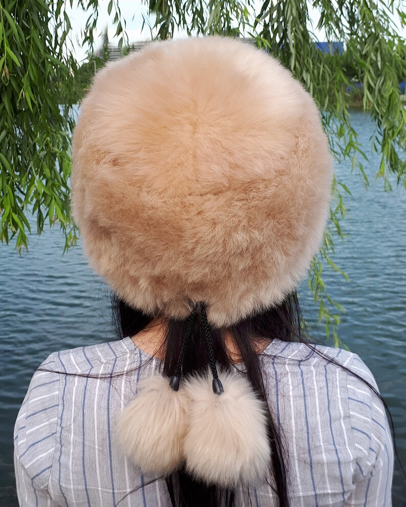 Peut inclure: Un bonnet rond en fausse fourrure beige, dot&eacute; d'une texture &eacute;paisse et douce. Le bonnet pr&eacute;sente deux pompons en fausse fourrure beige assortis, suspendus &agrave; de fines cordes noires &agrave; l'arri&egrave;re, formant un d&eacute;tail d&eacute;coratif sur le haut du dos.