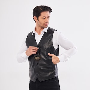 May include: A black leather vest featuring a V-neckline, three front buttons, and two horizontal zippered pockets at the waist. A small dark brown tag with the text DERI SITESI is attached to the front of the vest over the chest area.