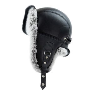 May include: A black leather trapper hat featuring thick, soft white and grey faux fur lining along the ear flaps and forehead. The hat includes a vertical black leather chin strap with two silver-toned metal snap buttons and a rectangular buckle.